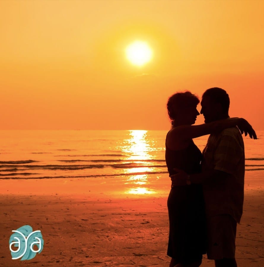 A silhouette of a couple embracing on the beach at sunset with orange skies and sea reflection.